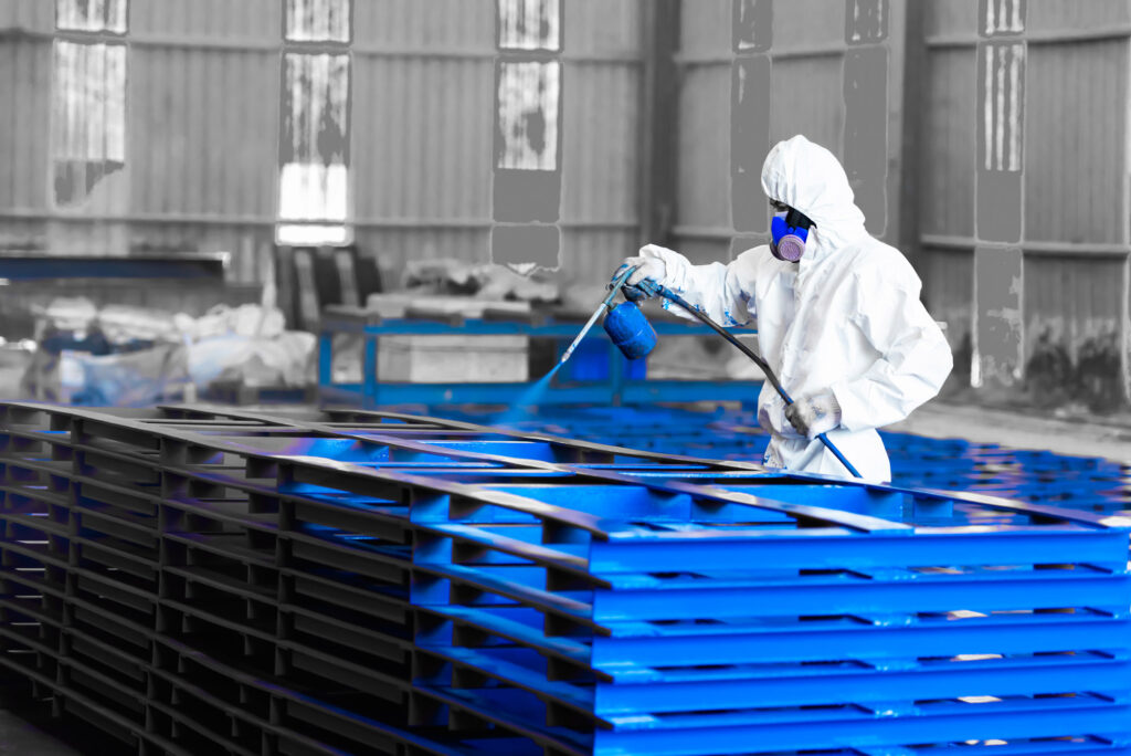 Industrial painter in protective gear applying blue coating to stacked steel beams in a factory setting
