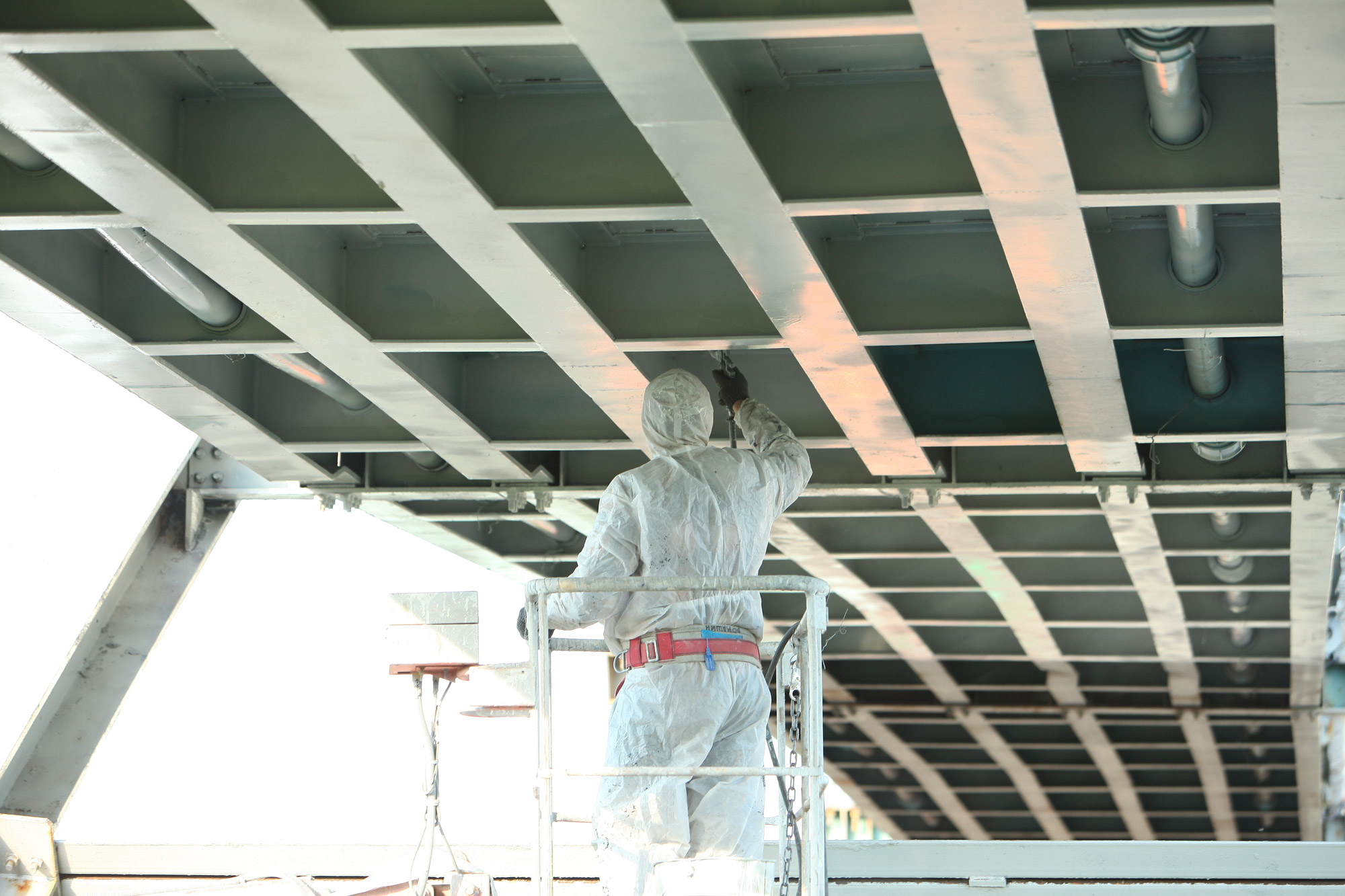 Industrial coating technician applying protective paint beneath a steel bridge using lift access